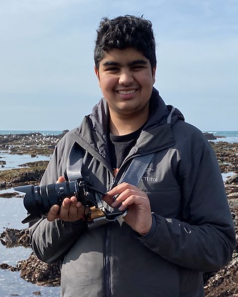 Devin Bhatt smiling on the San Francisco coast holding a mirroless camera.
